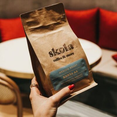 A hand holding a Skola coffee pack with a café setting in the background.